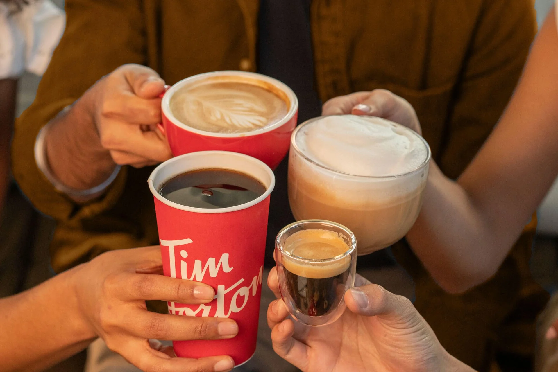 Three hands clinking Tim Hortons coffee cups in a cheers — a latte, a black coffee, and an espresso